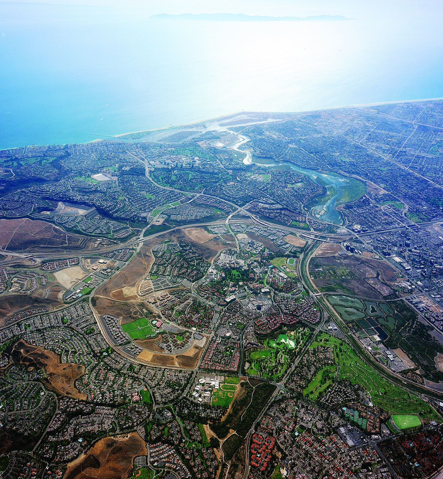 An aerial view of UC Irvine campus shows buildings surrounded by greenery and roads and the ocean in the distance